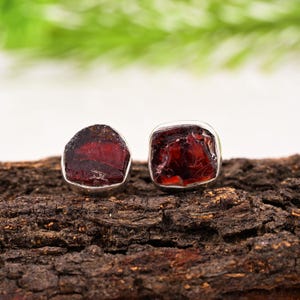 Pode incluir: Dois brincos de granada vermelha escura bruta, com engaste em prata. Os brincos s&atilde;o exibidos em um peda&ccedil;o de madeira escura e texturizada, com um fundo verde e branco desfocado.