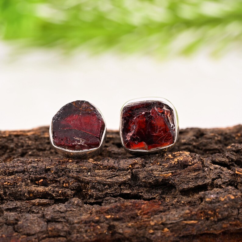 Pode incluir: Dois brincos de granada vermelha escura bruta, com engaste em prata. Os brincos s&atilde;o exibidos em um peda&ccedil;o de madeira escura e texturizada, com um fundo verde e branco desfocado.