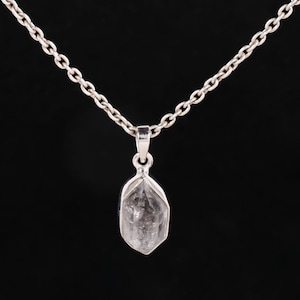 Pu&ograve; includere: Una collana d'argento con un ciondolo di cristallo di quarzo trasparente. Il ciondolo ha una forma sfaccettata e allungata, sospeso a una delicata catena. Lo sfondo &egrave; nero.