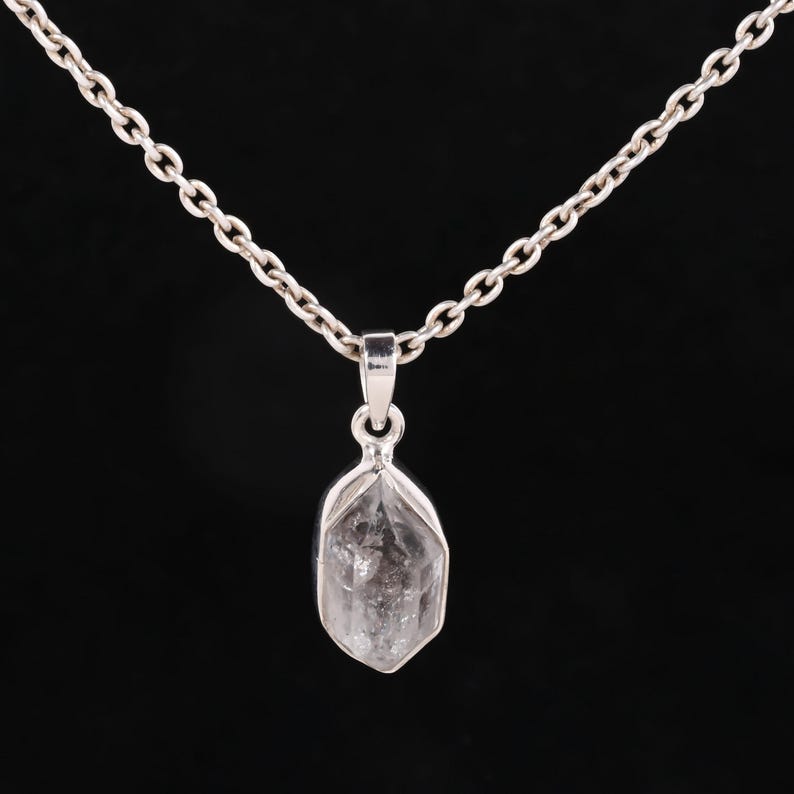 Pu&ograve; includere: Una collana d'argento con un ciondolo di cristallo di quarzo trasparente. Il ciondolo ha una forma sfaccettata e allungata, sospeso a una delicata catena. Lo sfondo &egrave; nero.