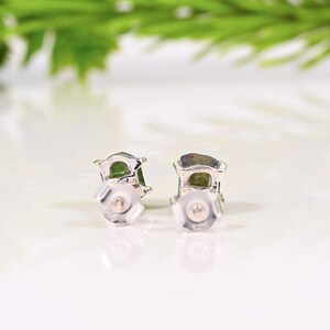 May include: A pair of silver stud earrings with green gemstones. The earrings are set in a silver frame and have a post and butterfly backing. The background is a blurred white surface with green foliage.