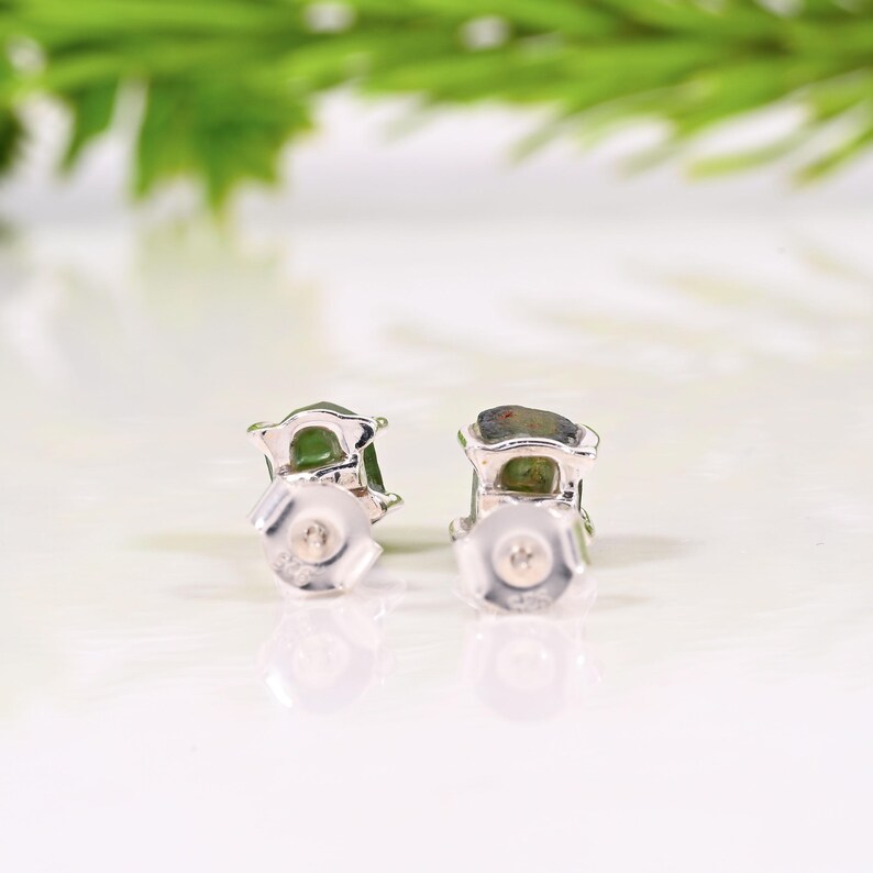 May include: A pair of silver stud earrings with green gemstones. The earrings are set in a silver frame and have a post and butterfly backing. The background is a blurred white surface with green foliage.