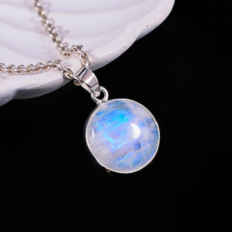 May include: A round, iridescent moonstone pendant necklace. The moonstone has a blue and white color scheme. The pendant is set in a silver-colored metal and hangs from a matching chain. The necklace is displayed against a dark background.