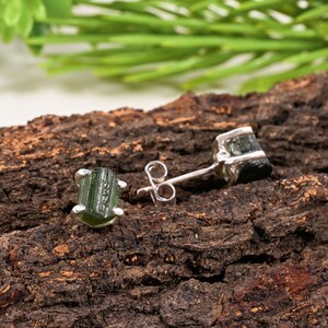 May include: A pair of green tourmaline stud earrings with a silver setting. The earrings are displayed on a textured, brown wooden surface. One earring is in focus, showing the gemstone's natural shape and color.