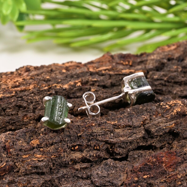 May include: A pair of green tourmaline stud earrings with a silver setting. The earrings are displayed on a textured, brown wooden surface. One earring is in focus, showing the gemstone's natural shape and color.
