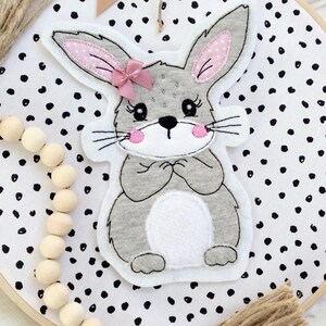 Puede incluir: Un conejito gris con orejas rosas y un lazo rosa está bordado en una tela blanca con lunares negros. La tela está estirada sobre un aro de bordado de madera. Una guirnalda de cuentas de madera cuelga del aro con un borla marrón.