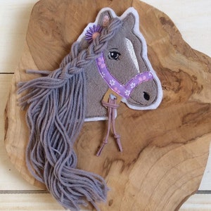May include: A grey horse head with a purple braided mane and a purple and white floral bridle. The horse head is a felt appliqué with a yarn mane.