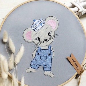 Puede incluir: Aro bordado gris con un lindo ratón de dibujos animados que lleva un gorro de marinero azul y blanco y un mono azul. El ratón está de pie sobre un fondo gris. El texto "Gracias" está escrito en una etiqueta marrón.