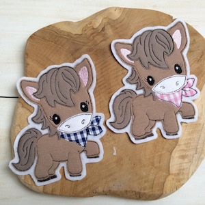 Può includere: Due applicazioni in feltro marrone di cavalli dei cartoni animati con fiocchi di nastro a quadri rosa e blu. I cavalli sono in piedi su una superficie di legno.