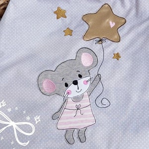 Puede incluir: Un saco de dormir gris con ribete blanco. El saco de dormir tiene un lindo diseño bordado de un ratón gris que lleva un vestido rosa y sostiene un globo de estrella dorada. El fondo es gris claro con lunares blancos.