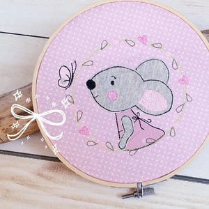 May include: A round embroidery hoop featuring a gray mouse wearing a pink dress, surrounded by a wreath of hearts and leaves. A black butterfly is near the mouse. The background is pink with white polka dots.