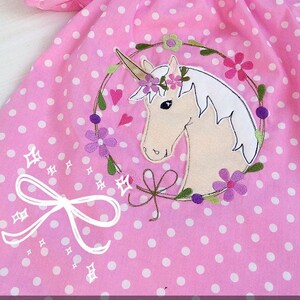 May include: Pink polka dot dress with a white unicorn embroidered on the front. The unicorn is wearing a flower crown and is surrounded by a wreath of flowers.