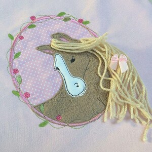 May include: A pink fabric applique of a brown horse head with a white nose and a pink flower wreath. The horse has a long, blonde mane.
