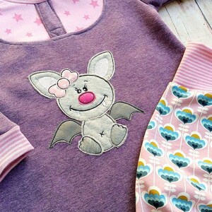 May include: Purple long-sleeved shirt with a pink and white star pattern on the top. The shirt features an embroidered grey bat with a pink bow and a pink nose. The shirt is paired with pink and white striped trousers with a floral pattern.