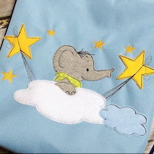 Puede incluir: Una bolsa de cordón azul claro con un diseño bordado de un elefante gris montando una nube blanca con estrellas amarillas. El elefante lleva una bufanda verde y blanca.