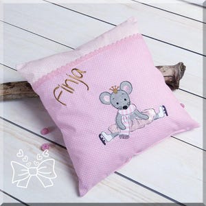 May include: Pink pillow with a white lace trim and a gray embroidered mouse wearing a crown and a pink tutu. The pillow has the name "Finja" embroidered on it.
