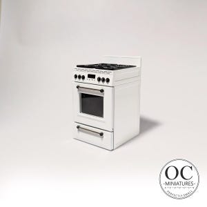 Puede incluir: Una estufa blanca en miniatura con una placa de cocina negra y una puerta de horno. La estufa tiene cuatro quemadores, perillas negras y un cajón. La puerta del horno tiene una ventana transparente. Es un modelo en miniatura.