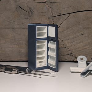 Könnte beinhalten: Ein Miniatur-Kühlschrank in Dunkelblau mit geöffneter Tür, der einen weißen Innenraum mit mehreren Regalen und einer unteren Schublade zeigt. Das Objekt steht neben einem Metallwerkzeug und einer weißen Büroklammer als Größenvergleich.