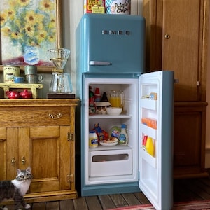 Könnte beinhalten: Ein hellblauer Miniatur-Kühlschrank der Marke Smeg mit geöffneter Tür, der mit winzigen Lebensmitteln wie einem Krug Orangensaft, Gewürzen und frischen Produkten gefüllt ist. Das Gerät hat einen silbernen Griff und ein Logo.