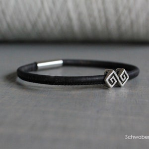 Könnte beinhalten: Ein schwarzes Korkarmband mit einem silbernen Anhänger mit einem doppelseitigen griechischen Schlüsselstil. Das Armband hat einen Schiebeverschluss.