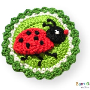 Pode incluir: Uma aplicação de joaninha de crochê com detalhes vermelhos e pretos, sentada em um círculo de crochê verde e branco.