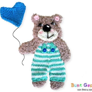May include: A handmade, knitted teddy bear with a blue heart-shaped balloon. The bear has brown fur, a white muzzle, and turquoise and white striped overalls. The balloon is a vibrant blue. The text "Bunt Gestrickt" is visible.
