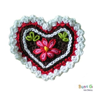 Könnte beinhalten: Ein gehäkeltes Herz-förmiges Ornament mit brauner Mitte und weißem Rand. Die Mitte zeigt eine rote Blume mit grünen Blättern.