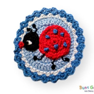 Pode incluir: Uma aplicação de joaninha de crochê com detalhes vermelhos e pretos sobre um fundo azul e branco. A joaninha tem um olho branco e uma boca vermelha.