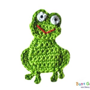 Könnte beinhalten: Ein grüner gehäkelter Frosch mit weißen Augen und einem roten Mund. Der Frosch sitzt auf einem weißen Hintergrund.
