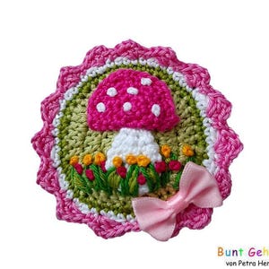 Pode incluir: Uma aplicação de cogumelo de crochê com detalhes em rosa e branco, cercada por folhas verdes e pequenas flores cor-de-rosa. Um laço rosa está preso à parte inferior da aplicação.