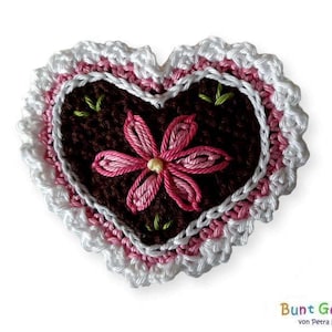 Könnte beinhalten: Eine gehäkelte Herz-Applikation mit einer pinkfarbenen Blume, die in der Mitte gestickt ist. Das Herz ist von einem weißen Häkelrand mit einem darunterliegenden pinkfarbenen Häkelrand umgeben.