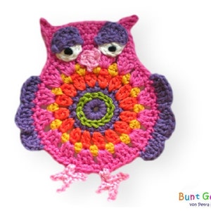 Op de afbeelding: Een gehaakte uil met een roze lichaam, paarse oren en een kleurrijk cirkelvormig patroon in het midden. De uil heeft zwarte ogen en een roze snavel.