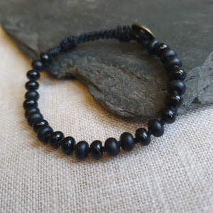 Puede incluir: Un brazalete negro de cuentas con un cordón negro y un pequeño acento dorado. El brazalete está sobre un fondo de tela marrón claro.