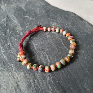 Può includere: Un braccialetto di corda rossa con perline colorate e macchiate. Il braccialetto è fatto di una corda rossa e ha una varietà di piccole perline rotonde in tonalità di rosso, arancione, giallo, verde, blu e viola.
