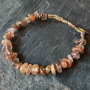 Könnte beinhalten: Ein Armband aus braunen und klaren Edelsteinen mit einem mehrfarbigen geflochtenen Band und einem goldfarbenen Verschluss.