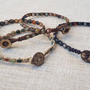 Peut inclure: Trois bracelets avec des cordons bruns, beiges et noirs, chacun avec un motif différent de pierres vertes, brunes et orange. Chaque bracelet a une fermeture bouton en bois.