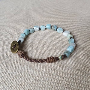 Może przedstawiać: Ręcznie robiona bransoletka z jasnoniebieskimi i białymi koralikami z kamieni szlachetnych. Bransoletka jest nawleczona na ciemnobrązowy sznurek z ozdobnym węzłem i mosiężnym zapięciem na guzik. Koraliki mają nieregularny kształt.