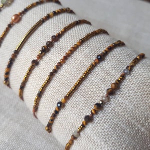 Può includere: Un set di sette braccialetti realizzati con perline di occhio di tigre in tonalità di marrone e oro. I braccialetti sono disposti in fila su uno sfondo di tessuto beige.