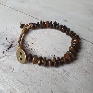 Könnte beinhalten: Ein braunes und goldenes Perlenarmband mit einem Knopfverschluss. Die Perlen sind aus Naturstein und auf einer dünnen Schnur aufgefädelt.