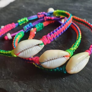 Puede incluir: Primer plano de tres pulseras de macramé coloridas. Cada pulsera presenta una concha blanca y un cordón de macramé de colores del arcoíris. Las pulseras son en tonos de rosa, verde, morado y naranja. Las pulseras son de estilo bohemio.