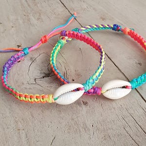 May include: Two colorful woven bracelets with a seashell accent. The bracelets feature a rainbow of colors, including pink, yellow, green, and blue. The seashells are white. The bracelets are on a light wood surface.