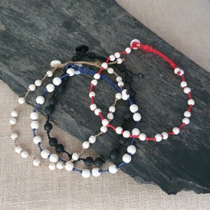 Può includere: Quattro braccialetti di perline con perline di lava bianche e nere. I braccialetti sono infilati su cordoncini di vari colori, tra cui rosso, blu e nero. I braccialetti sono esposti su una superficie strutturata grigio scuro.