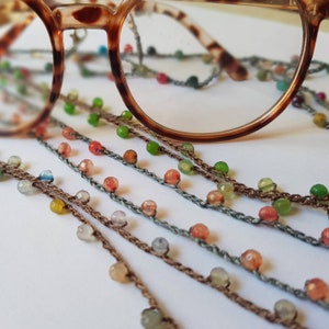 Peut inclure: Gros plan sur plusieurs fils de colliers de perles avec des perles de différentes couleurs, notamment vertes, rouges et bleues. Les colliers sont faits d'un cordon fin crocheté. Une paire de lunettes en écaille de tortue est posée sur les colliers.