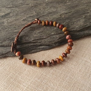 Puede incluir: Un brazalete de cordón de cuero marrón con una cadena de cuentas de jaspe multicolores. Las cuentas son una mezcla de rojo, naranja, amarillo y marrón.