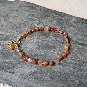 Puede incluir: Un brazalete de oro con cuentas marrones, naranjas y blancas. El brazalete tiene un cierre de oro y un pequeño dije de oro.