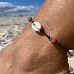 Peut inclure: Un bracelet de cheville marron avec des coquillages blancs et des perles colorées. Le bracelet est porté à la cheville d'une personne.