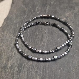 Peut inclure: Deux bracelets de perles argentées avec un mélange de perles carrées et rondes. Les bracelets sont reliés par un fermoir noir. Les bijoux sont présentés sur une surface en pierre grise.