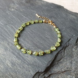 Puede incluir: Un delicado brazalete de piedras preciosas verdes con un cierre de oro. El brazalete está hecho de pequeñas piedras verdes de forma irregular enhebradas en una fina cadena de oro.