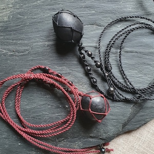 Può includere: Due ciondoli in pietra nera con un motivo geometrico, ognuno appeso a un cordoncino. Un cordoncino è nero e l'altro è bordeaux.