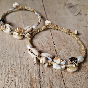 Puede incluir: Dos pulseras trenzadas adornadas con conchas y perlas. Una pulsera presenta una trenza plateada y beige, mientras que la otra tiene una trenza dorada. Las conchas son en tonos beige, blanco y marrón.
